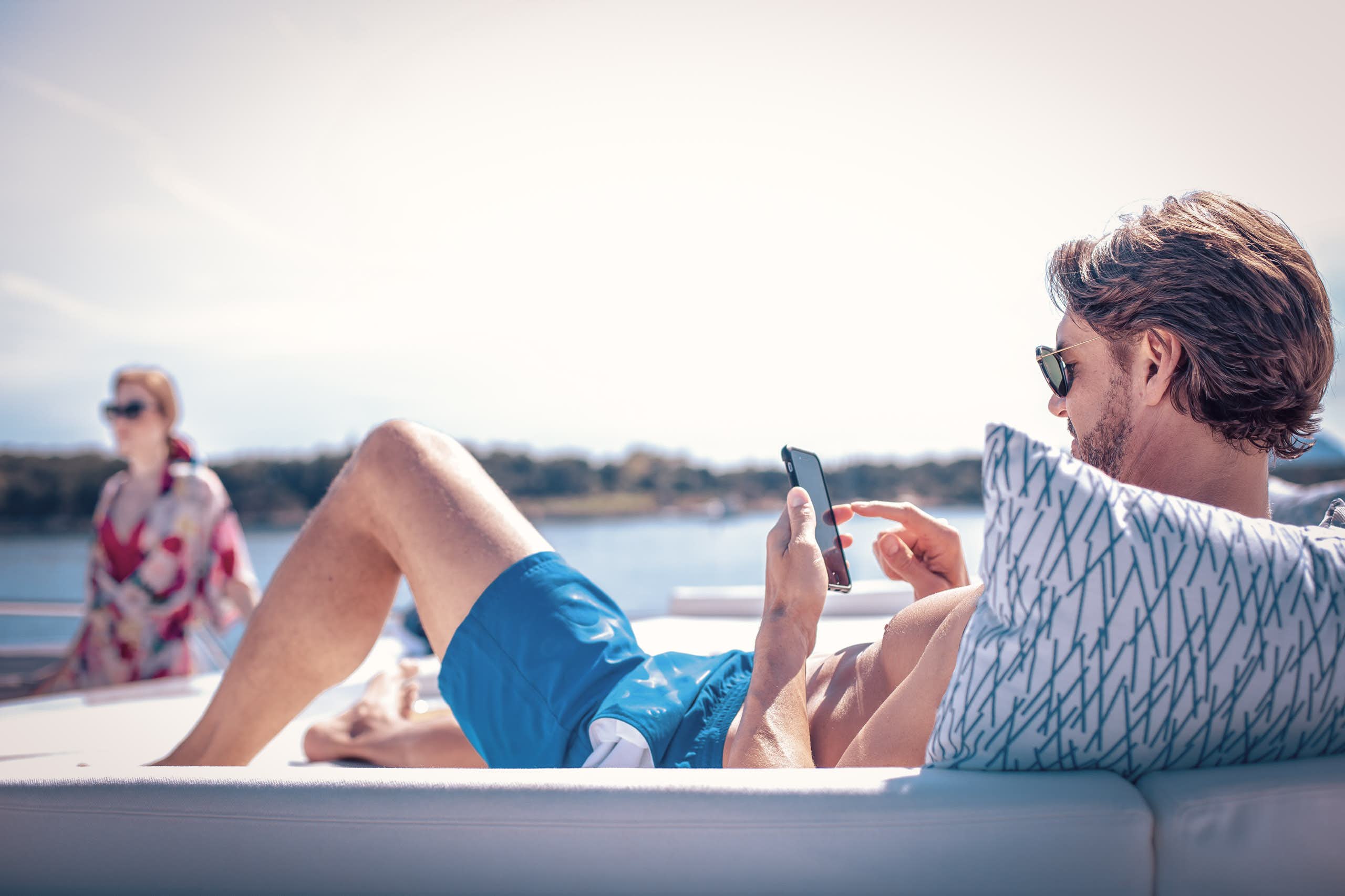 Man looking at mobile device while on sundeck with woman on background